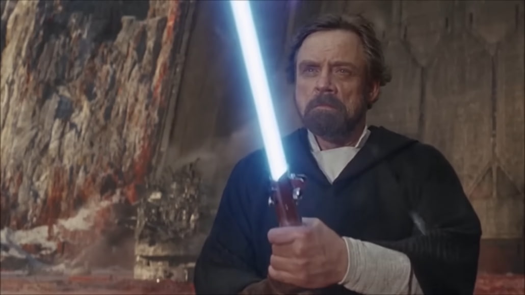 Star Wars character standing with a blue sword up, rocks behind him